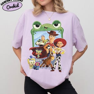 May include: A light purple t-shirt featuring a graphic print of Toy Story 5 characters including Woody, Buzz Lightyear, Jessie, and Bullseye. The design includes a green frog frame, the Toy Story 5 logo, and text stating in theaters June 19.