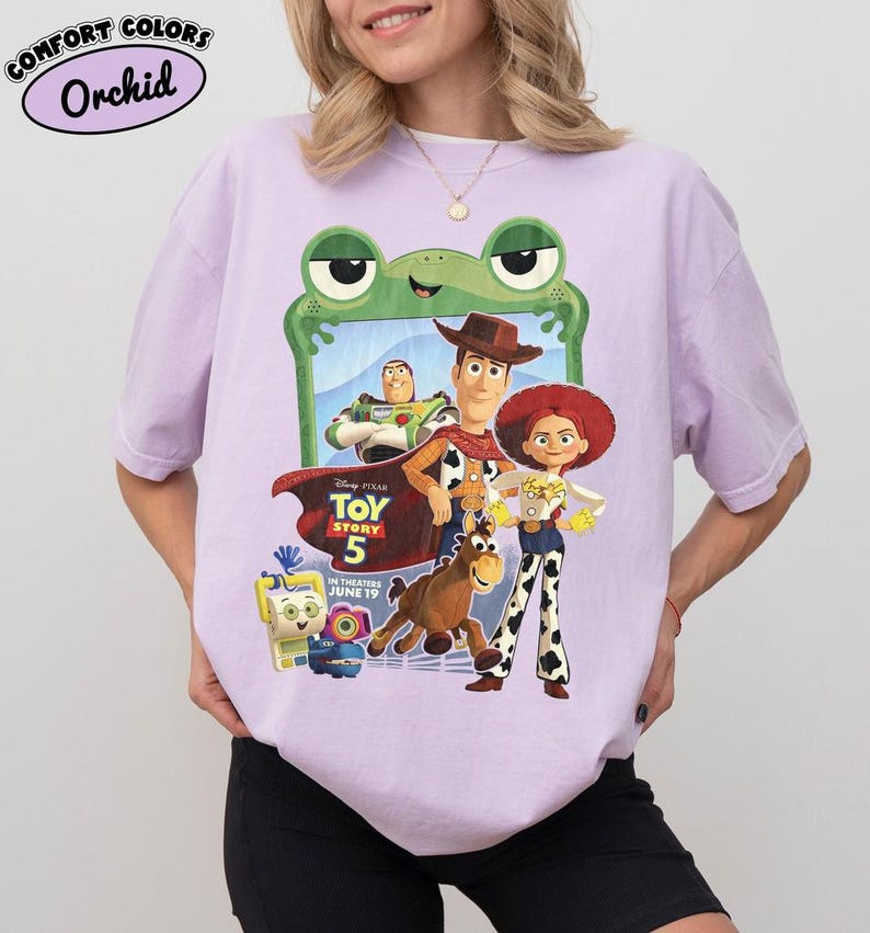 May include: A light purple t-shirt featuring a graphic print of Toy Story 5 characters including Woody, Buzz Lightyear, Jessie, and Bullseye. The design includes a green frog frame, the Toy Story 5 logo, and text stating in theaters June 19.