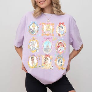 May include: A light purple t-shirt featuring nine framed portraits of Disney princesses. Each portrait is surrounded by floral and bow accents. The princesses are depicted in their iconic outfits and hairstyles. The shirt is a comfortable, casual style.