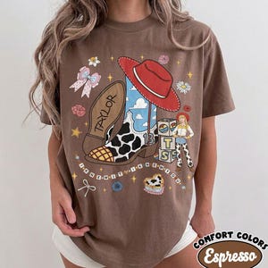 May include: A brown t-shirt featuring a graphic print of a cowboy boot with a cow print pattern, a red cowboy hat, a cartoon character, and alphabet blocks. The text "TAYLOR" and "I KNEW IT I KNEW YOU" are printed alongside stars, flowers, and a bow.