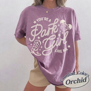 Op de afbeelding: Een paars T-shirt met korte mouwen en een cr&egrave;mekleurige opdruk met de tekst "YOU'RE A Park Girl LIKE ME". Het ontwerp bevat een cowboylaars, sterren en twee strikken. Het T-shirt heeft een ronde hals en een losvallend silhouet.