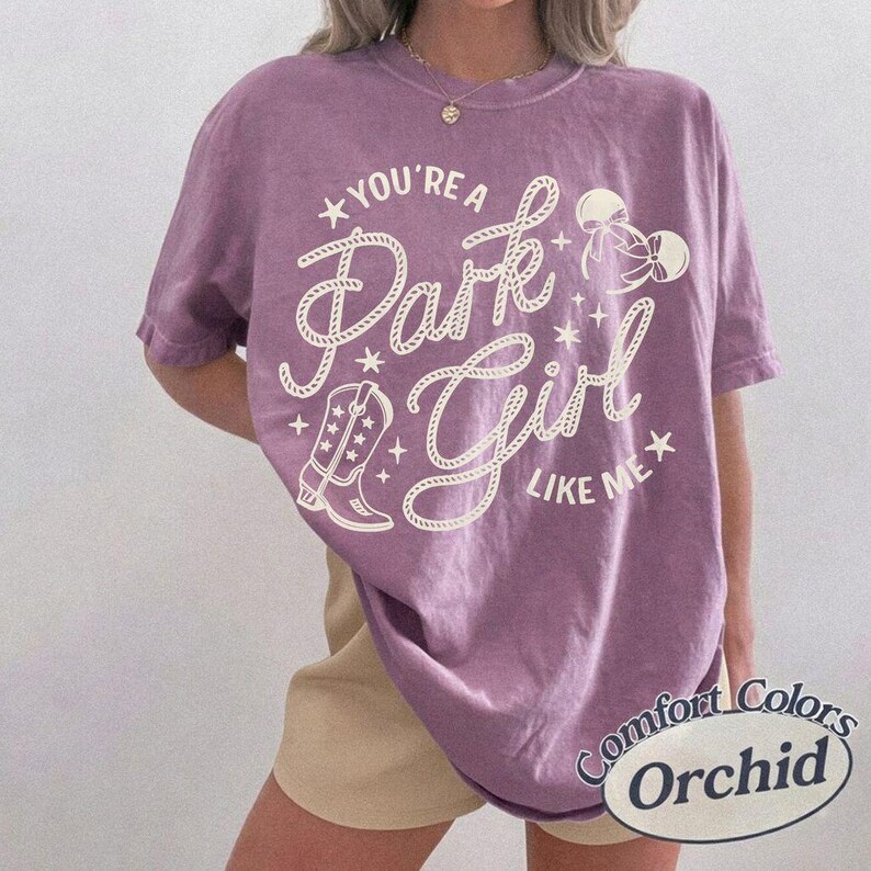 Op de afbeelding: Een paars T-shirt met korte mouwen en een cr&egrave;mekleurige opdruk met de tekst "YOU'RE A Park Girl LIKE ME". Het ontwerp bevat een cowboylaars, sterren en twee strikken. Het T-shirt heeft een ronde hals en een losvallend silhouet.