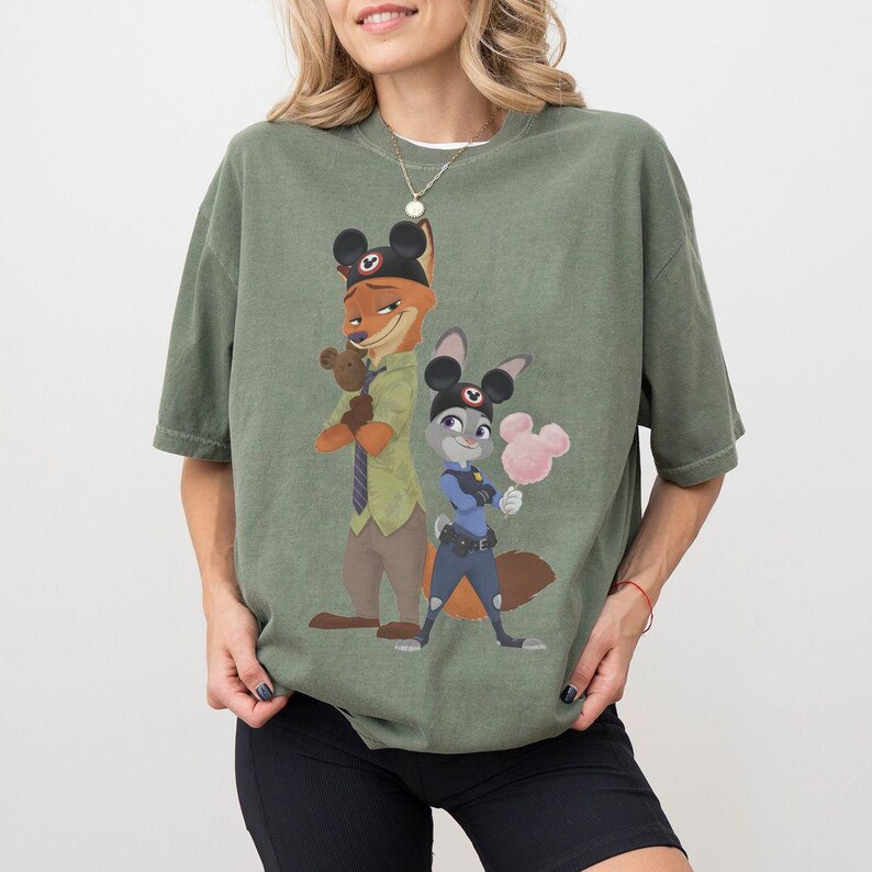 Pu&ograve; includere: T-shirt verde oliva con un'illustrazione di Nick Wilde e Judy Hopps di Zootopia, ognuno con le orecchie di Topolino. Nick tiene un orsacchiotto, e Judy uno zucchero filato rosa. La maglietta &egrave; di vestibilit&agrave; ampia.