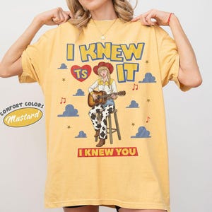 May include: A mustard yellow t-shirt featuring a graphic of a person in a cowboy hat and cow-print pants playing an acoustic guitar on a stool. The text reads I KNEW TS IT and I KNEW YOU, surrounded by red hearts, blue clouds, and gold stars.
