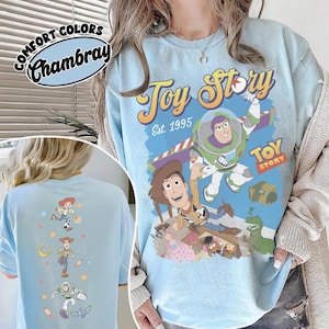 May include: Light blue t-shirt featuring the text "Toy Story Est. 1995" and images of Woody and Buzz Lightyear. The back of the shirt has images of Jessie, Woody, and Buzz Lightyear. The shirt is labeled "Comfort Colors Chambray".