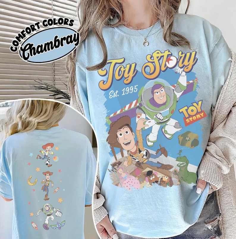 May include: Light blue t-shirt featuring the text "Toy Story Est. 1995" and images of Woody and Buzz Lightyear. The back of the shirt has images of Jessie, Woody, and Buzz Lightyear. The shirt is labeled "Comfort Colors Chambray".
