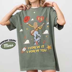 Puede incluir: Una camiseta verde musgo con un gr&aacute;fico de una persona de pelo largo tocando la guitarra, con una camisa de cuadros amarillos y pantalones con estampado de vaca. El dise&ntilde;o incluye un sombrero rojo, un coraz&oacute;n con TS, un cohete, estrellas y el texto I KNEW IT I KNEW YOU.