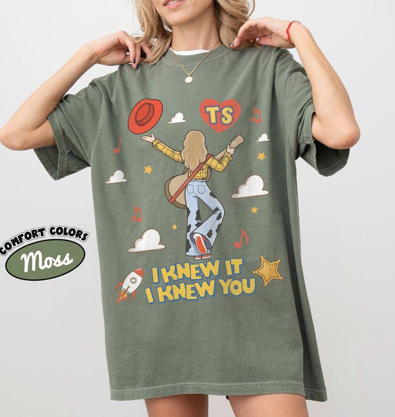 Puede incluir: Una camiseta verde musgo con un gr&aacute;fico de una persona de pelo largo tocando la guitarra, con una camisa de cuadros amarillos y pantalones con estampado de vaca. El dise&ntilde;o incluye un sombrero rojo, un coraz&oacute;n con TS, un cohete, estrellas y el texto I KNEW IT I KNEW YOU.