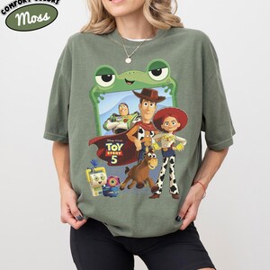 May include: A moss green t-shirt featuring a graphic print of Toy Story 5 characters including Woody, Jessie, Buzz Lightyear, Bullseye, and others framed by a green frog outline. The shirt includes the Toy Story 5 logo and Comfort Colors branding.
