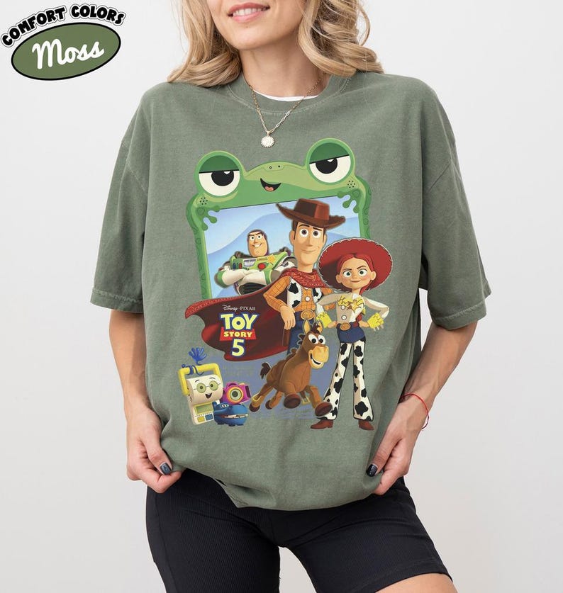May include: A moss green t-shirt featuring a graphic print of Toy Story 5 characters including Woody, Jessie, Buzz Lightyear, Bullseye, and others framed by a green frog outline. The shirt includes the Toy Story 5 logo and Comfort Colors branding.