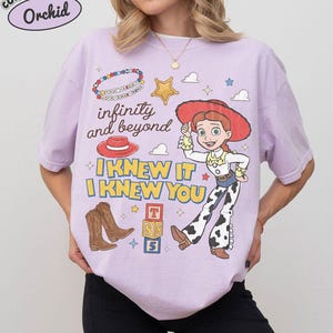May include: A light purple t-shirt featuring a graphic print of a cowgirl character, a red hat, brown cowboy boots, a gold star, and alphabet blocks. The text reads "infinity and beyond" and "I KNEW IT I KNEW YOU" with a beaded bracelet graphic.