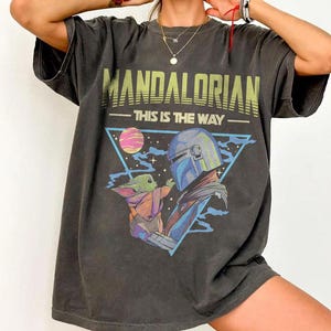 Może przedstawiać: Ciemnoszary t-shirt z nadrukiem graficznym zawierającym napis MANDALORIAN oraz THIS IS THE WAY. Wz&oacute;r przedstawia postać w niebieskim hełmie i małe zielone stworzenie wewnątrz niebieskiego tr&oacute;jkąta, na tle r&oacute;żowo-fioletowej planety.