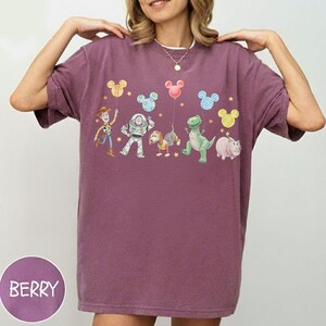 May include: Berry-colored t-shirt featuring a graphic of animated characters and Mickey Mouse-shaped balloons. The design includes Woody, Buzz Lightyear, Rex, Hamm, and Slinky Dog. The shirt has a relaxed fit and short sleeves.