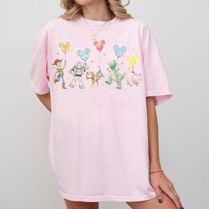 May include: A light pink t-shirt featuring a colorful graphic of cartoon characters and balloons. The design includes characters like Woody, Buzz Lightyear, and Rex. The balloons are in the shape of Mickey Mouse heads.