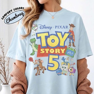 Puede incluir: Una camiseta azul claro con un estampado gr&aacute;fico de los personajes de Disney Pixar Toy Story 5, incluyendo a Woody, Buzz Lightyear, Rex, Jessie, Bullseye y Forky. El texto dice Disney Pixar Toy Story 5 con un logo Comfort Colors Chambray.