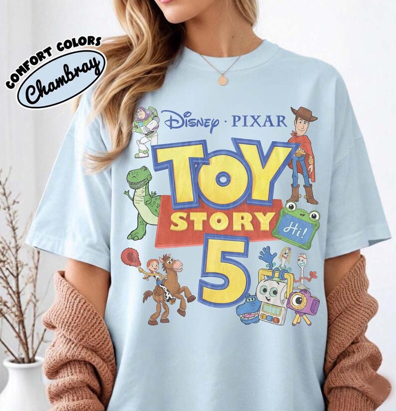 Puede incluir: Una camiseta azul claro con un estampado gr&aacute;fico de los personajes de Disney Pixar Toy Story 5, incluyendo a Woody, Buzz Lightyear, Rex, Jessie, Bullseye y Forky. El texto dice Disney Pixar Toy Story 5 con un logo Comfort Colors Chambray.