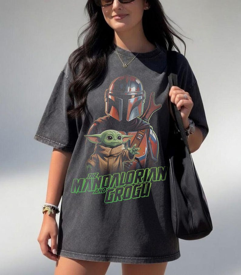 May include: A charcoal grey oversized t-shirt featuring a graphic print of The Mandalorian and Grogu. The design includes the characters in their signature armor and robes, with the text THE MANDALORIAN AND GROGU printed in bright green.