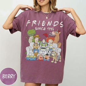 K&ouml;nnte beinhalten: Beerenfarbenes T-Shirt mit einer Grafik von Toy Story-Charakteren in einem Friends-Themen-Design. Der Text "FRIENDS SINCE 1995" ist &uuml;ber den Charakteren aufgedruckt. Das Shirt hat kurze &Auml;rmel und eine lockere Passform.