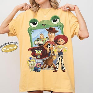 May include: A mustard yellow t-shirt featuring a graphic print of Toy Story 5 characters including Woody, Buzz Lightyear, Jessie, and Bullseye framed by a green frog outline. The text reads Disney Pixar Toy Story 5 In Theaters June 19.