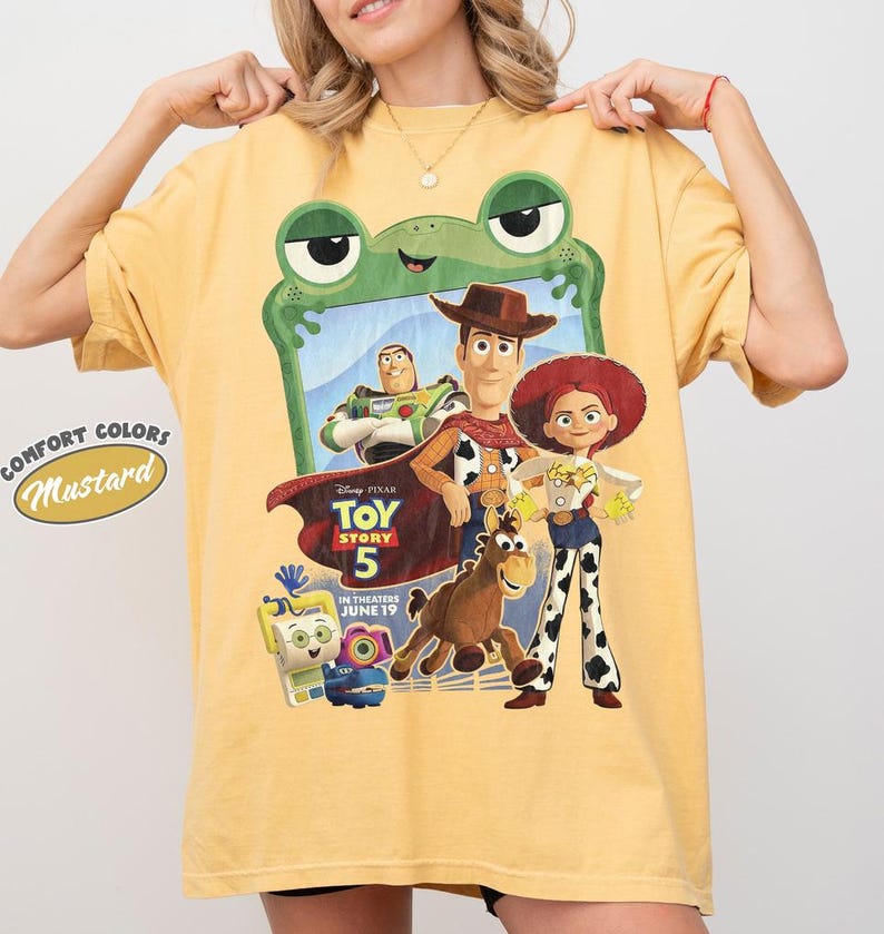 May include: A mustard yellow t-shirt featuring a graphic print of Toy Story 5 characters including Woody, Buzz Lightyear, Jessie, and Bullseye framed by a green frog outline. The text reads Disney Pixar Toy Story 5 In Theaters June 19.