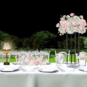 Può includere: Una scena di cena all'aperto splendidamente allestita con una lunga tovaglia bianca, sedie bianche e composizioni floreali rosa e bianche. Un lampadario di cristallo e alti centrotavola floreali aggiungono eleganza.