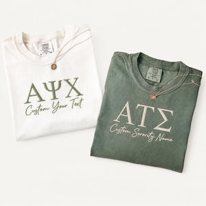 May include: A white t-shirt with olive green Greek letters and the text "Custom Your Text" and a sage green t-shirt with white Greek letters and the text "Custom Sorority Name," each featuring a delicate gold-toned necklace resting on the collar.