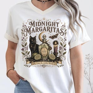 May include: A white V-neck t-shirt featuring a graphic print with the text "MIDNIGHT MARGARITAS" and "Salt, Lime & a Little Witchcraft". The design includes a black cat, a crescent moon, a moth, a candle, and a bottle labeled "LUNA AGAVE ELIXIR".