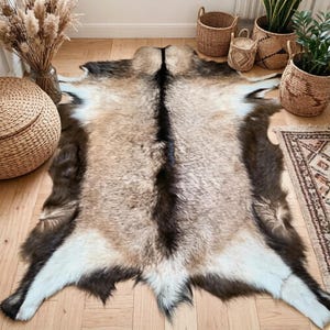 Puede incluir: Una alfombra de piel de animal de tonos naturales con una franja marrón oscuro en el centro. La alfombra presenta una mezcla de pelo marrón claro, marrón oscuro y blanco. La alfombra se exhibe sobre un suelo de madera, con cestas decorativas y plantas en el fondo.