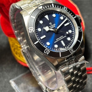Puede incluir: Un reloj de pulsera de acero inoxidable con esfera azul y bisel negro. El reloj presenta el texto "TUDOR GENEVE" y "200m:660ft MASTER CHRONOMETER". La correa es de acero inoxidable.