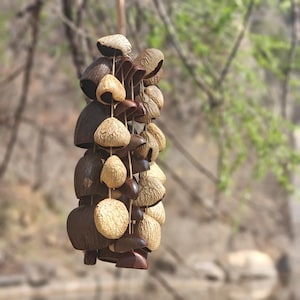 Puede incluir: Un m&oacute;vil colgante con una colecci&oacute;n de elementos de madera natural, texturizada, marr&oacute;n y beige. El m&oacute;vil incluye varias formas, incluyendo formas de campana y piezas redondeadas, ensartadas con cuerdas finas. El fondo est&aacute; borroso, sugiriendo un entorno al aire libre.