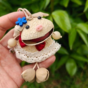 以下が含まれることがあります： ベージュの体、ピンクの鼻、黒い目をした手作りの豚のオーナメント。赤い蝶ネクタイ、青い花、白いレースの襟が付いています。豚には、ベージュのビーズが付いたぶら下がった脚と腕があります。