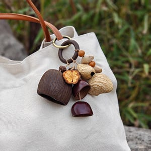 May include: A beige tote bag with brown leather handles and a decorative charm. The charm includes a wooden ring, beads, dried fruit, and seed pods in brown and beige tones.