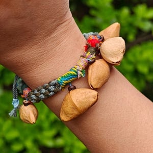 Puede incluir: Una pulsera hecha a mano con una banda trenzada en tonos verdes, azules y amarillos, adornada con vainas de semillas naturales de color marrón claro. La pulsera tiene una estética rústica y bohemia, con acentos de hilo colorido.