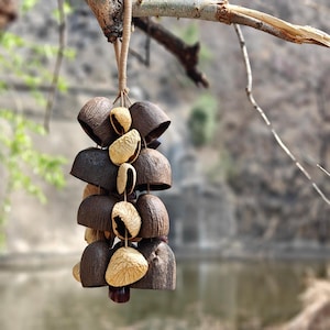 Puede incluir: Un carill&oacute;n colgante hecho de materiales naturales. El carill&oacute;n presenta piezas oscuras en forma de campana y vainas de semillas m&aacute;s claras, unidas con un cord&oacute;n tejido. El fondo borroso muestra un entorno natural al aire libre.