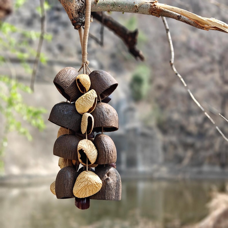 Puede incluir: Un carill&oacute;n colgante hecho de materiales naturales. El carill&oacute;n presenta piezas oscuras en forma de campana y vainas de semillas m&aacute;s claras, unidas con un cord&oacute;n tejido. El fondo borroso muestra un entorno natural al aire libre.