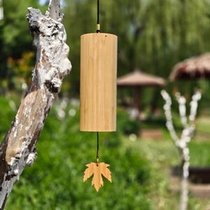 Könnte beinhalten: Ein zylindrisches Windspiel aus Bambus mit einer hellbraunen Holzmaserung. Eine schwarze Schnur hält den Hauptkörper an einer oberen Befestigung, während ein kleineres, blattförmiges Holzstück an einer dünnen schwarzen Schnur unten hängt.