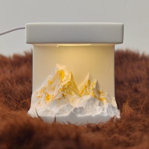 May include: A white, rectangular night light featuring a mountain range design. The mountains are white with gold detailing, and the light gives off a warm glow. The night light is plugged in with a white cable.