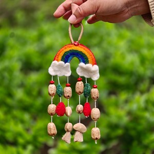 May include: A colorful rainbow-shaped wall hanging with a felted design. The rainbow features vibrant bands of red, orange, yellow, green, and blue. White felt clouds are attached below, with hanging beads in various colors and shapes. A decorative piece.