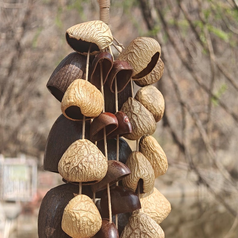 Puede incluir: Un carill&oacute;n de viento colgante con elementos naturales. Incluye piezas en forma de campana y vaina en tonos marrones y beige, unidas con cuerda. El dise&ntilde;o evoca una est&eacute;tica r&uacute;stica o bohemia, ideal para exteriores.
