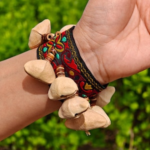 Puede incluir: Una pulsera con una banda negra, bordados rojos y dorados, y varias vainas de semillas grandes y de color claro. La pulsera se lleva en una muñeca, con un fondo verde.