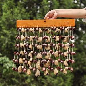 Puede incluir: Un carillón de viento de madera con una parte superior rectangular y múltiples hilos de cuentas de color natural y objetos marrones en forma de campana. El carillón de viento está sostenido por una mano sobre un fondo de follaje verde.