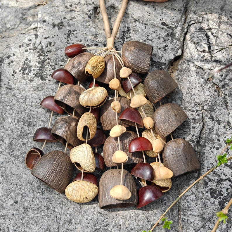 Puede incluir: Un collar artesanal hecho con elementos naturales. Presenta un conjunto de cuentas de madera y vainas de semillas en tonos marr&oacute;n, beige y burdeos, ensartadas con un cord&oacute;n natural, creando un dise&ntilde;o &uacute;nico y org&aacute;nico.