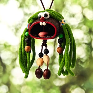 Può includere: Un ornamento sospeso realizzato con un guscio di noce di cocco marrone scuro con una bocca larga bordata di rosso, due denti bianchi e una lingua rosa. Presenta lunghi capelli in filato verde, perline di legno e semi chiari su cordini.