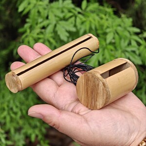 May include: Two bamboo musical instruments. One is cylindrical with a long slit, the other is shorter with a rectangular opening. Both are light brown with a black string attached. The background is blurred green foliage.