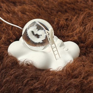 以下が含まれることがあります： 白い渦巻き模様が内側にある透明なガラス球が、白い雲型の台座の上に置かれています。梯子を持ったミニチュアの人物が球に向かって登っています。台座は茶色のテクスチャのある表面にあります。