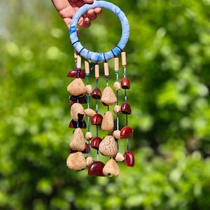 Peut inclure: Un carillon éolien artisanal avec un anneau supérieur recouvert de tissu bleu. Des brins de gousses texturées de couleur naturelle et d'éléments en forme de cloche rouge foncé pendent en dessous. Des perles de bois et des accents colorés sont intercalés.