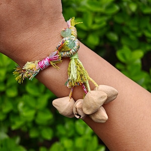 May include: A handmade bracelet featuring a woven design with colorful threads in pink, yellow, and green. The bracelet includes a green bead and several light brown seed pods, creating a unique, natural aesthetic.