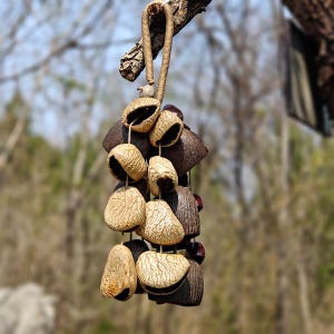Puede incluir: Un m&oacute;vil colgante con elementos naturales y org&aacute;nicos. El m&oacute;vil presenta una serie de vainas beige texturizadas y formas geom&eacute;tricas de color marr&oacute;n oscuro, unidas con una cuerda trenzada. Cuelga de una rama de &aacute;rbol.