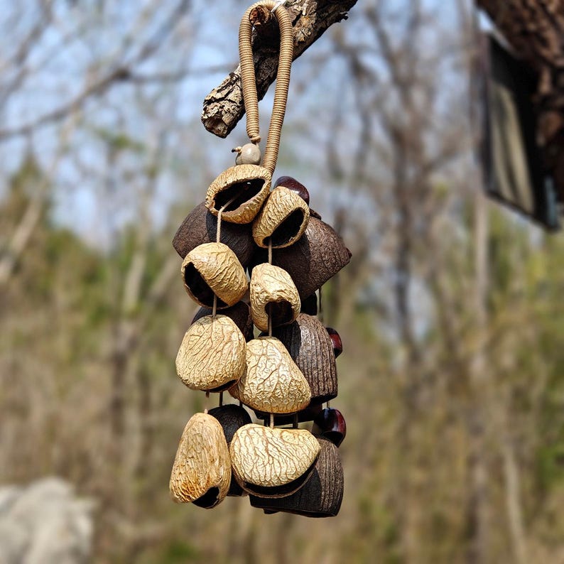 Puede incluir: Un m&oacute;vil colgante con elementos naturales y org&aacute;nicos. El m&oacute;vil presenta una serie de vainas beige texturizadas y formas geom&eacute;tricas de color marr&oacute;n oscuro, unidas con una cuerda trenzada. Cuelga de una rama de &aacute;rbol.