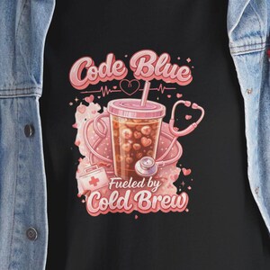 May include: Black t-shirt with a graphic design featuring the words "Code Blue" and "Fueled by Cold Brew." The design includes a coffee drink, stethoscope, and medical symbols in pink and white.